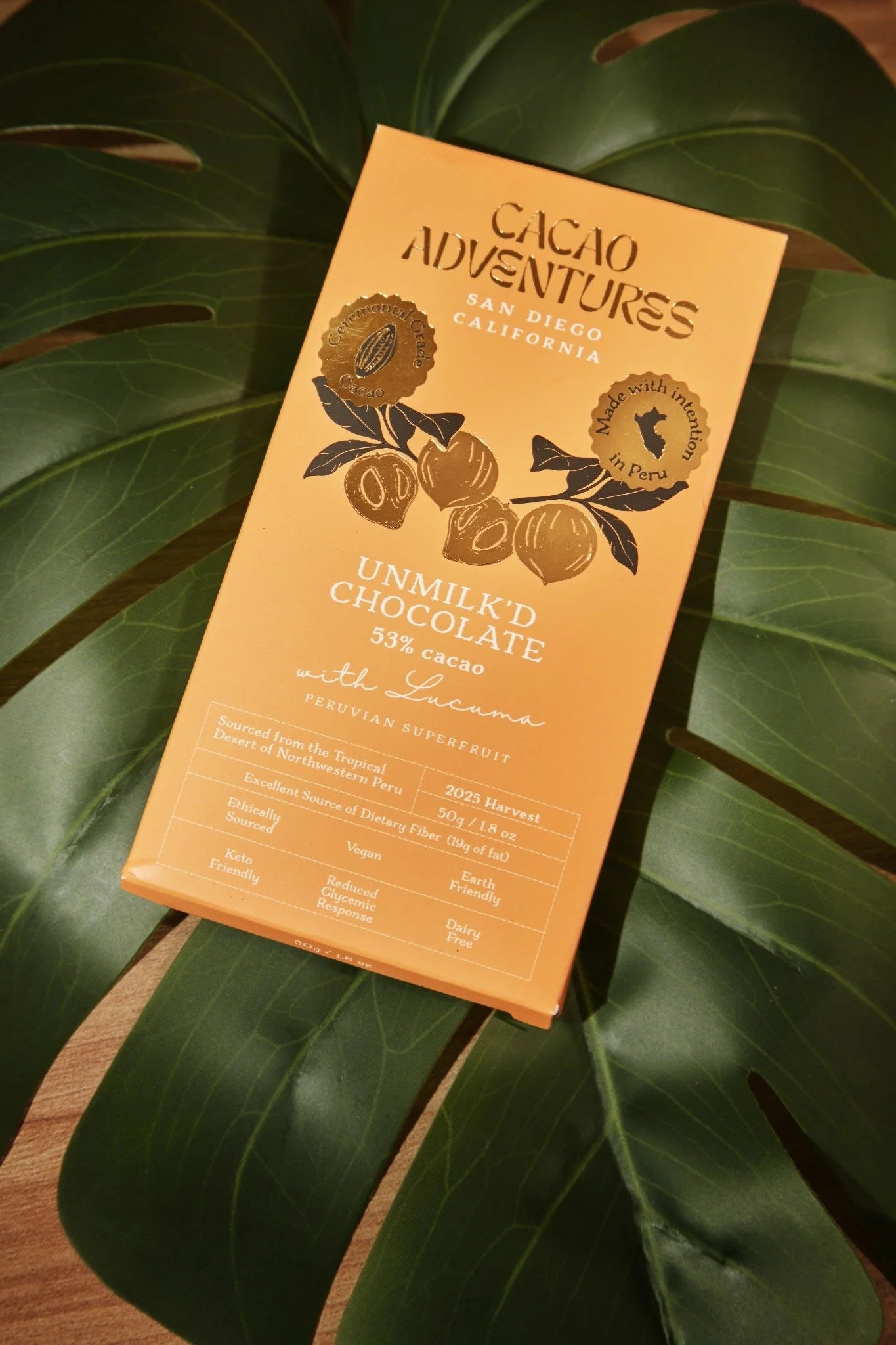 A rectangular box of Cacao Adventures 53% cocoa with Licorice chocolate, placed diagonally on large green monstera leaves over a wooden surface.
