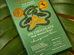  70% Chocolate Bar packaging from the High Amazon Basin, photographed at an angle on large green leaves. The package features a snake wrapping around cocoa pods and includes text such as 'Made with intention in Peru'.