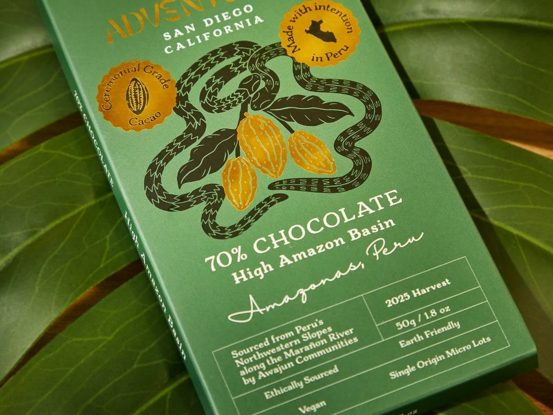  70% Chocolate Bar packaging from the High Amazon Basin, photographed at an angle on large green leaves. The package features a snake wrapping around cocoa pods and includes text such as 'Made with intention in Peru'.
