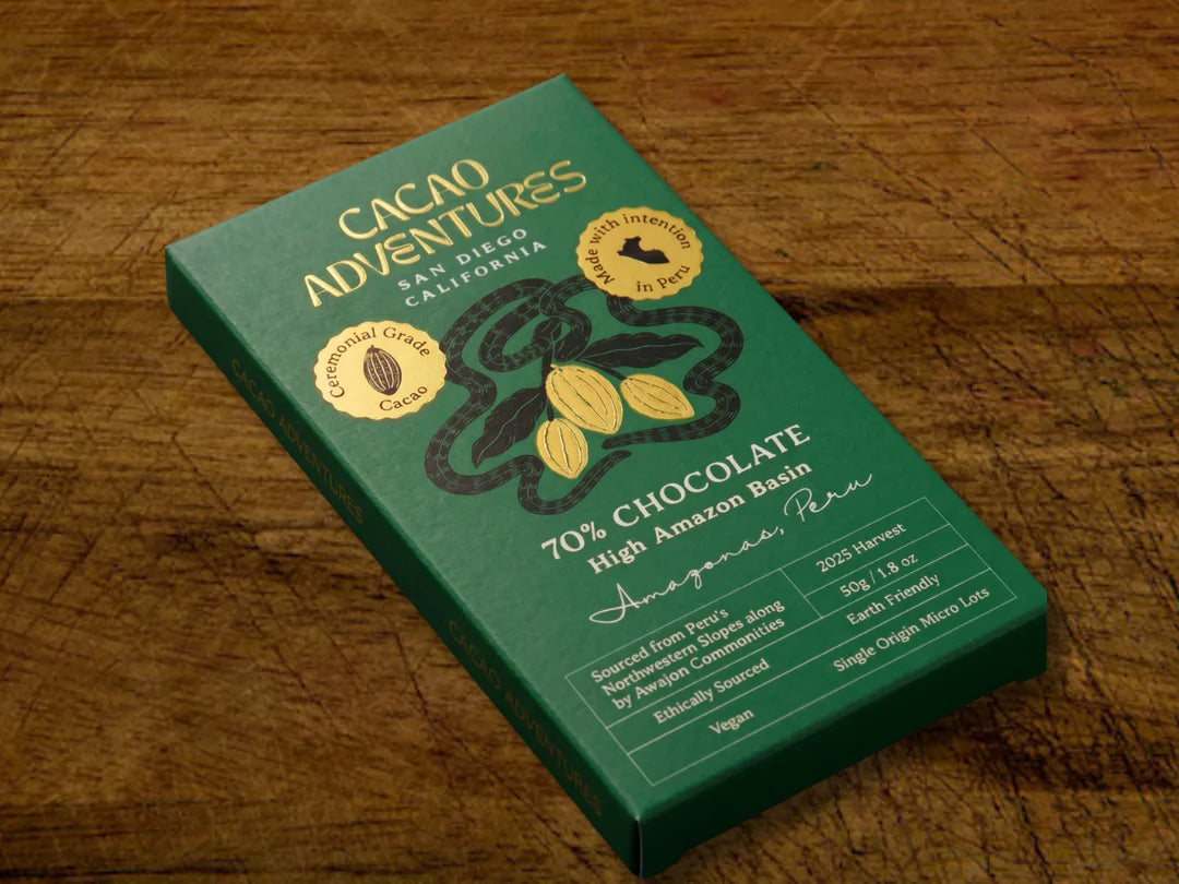 A green bar of Cacao Adventures chocolate placed on a rustic wooden table. The packaging features gold and white text, a cocoa bean illustration, and circular badges indicating ceremonial grade cacao and Made with Intention in Peru.