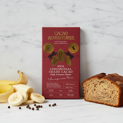 Cacao Adventures 100% Ceremonial Grade Cacao High Amazon Basin bar in burgundy and gold packaging standing upright on white marble, styled with fresh banana, allspice berries, and a slice of banana bread