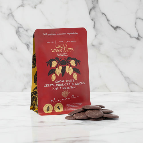 Cacao Adventures Ceremonial Cacao Paste red pouch standing upright with dark cacao paste discs in foreground on marble background, High Amazon Peru