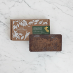 Untemptered 100% Cacao Paste Brick next to a rustic, textured dark brown wrapper in front of its packaging box on a marble background. The box is brown with white illustrations of monkeys and cocoa plants.