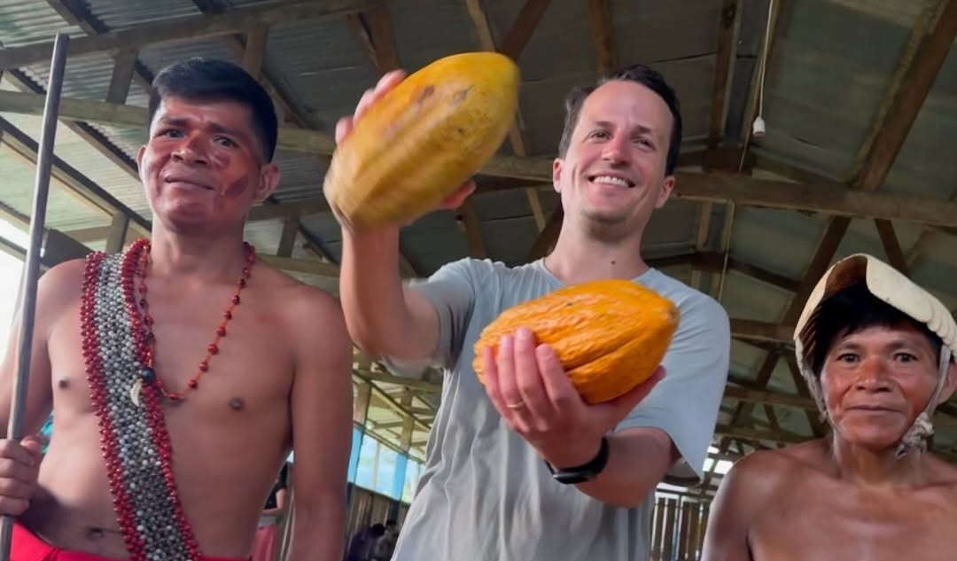 A smiling man in a gray t-shirt holds two yellow-orange cacao pods in a rustic indoor setting, flanked by two shirtless local men wearing red cloth and bead necklaces.
