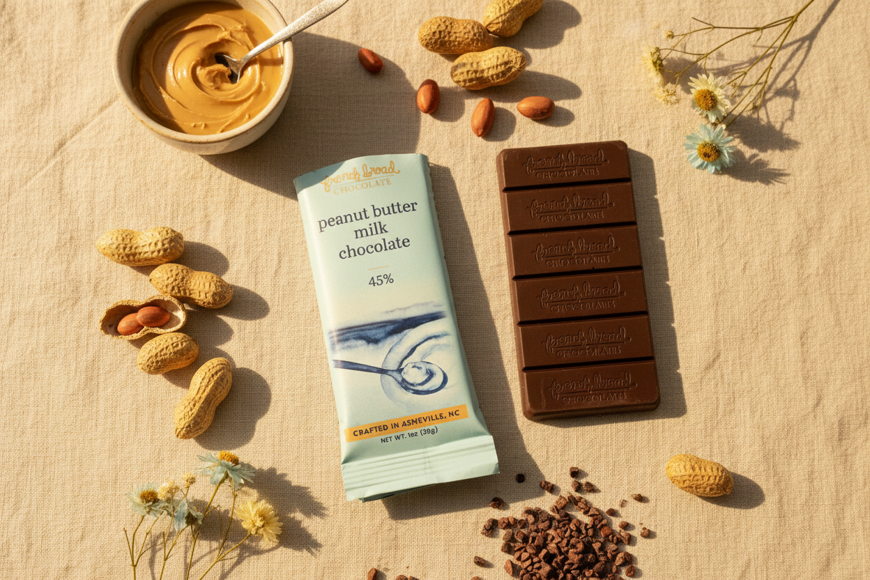 French Broad Chocolate 45% Peanut Butter Milk Chocolate bar and packaging flat lay with peanut butter bowl, cacao nibs, peanuts, and wildflowers on linen surface