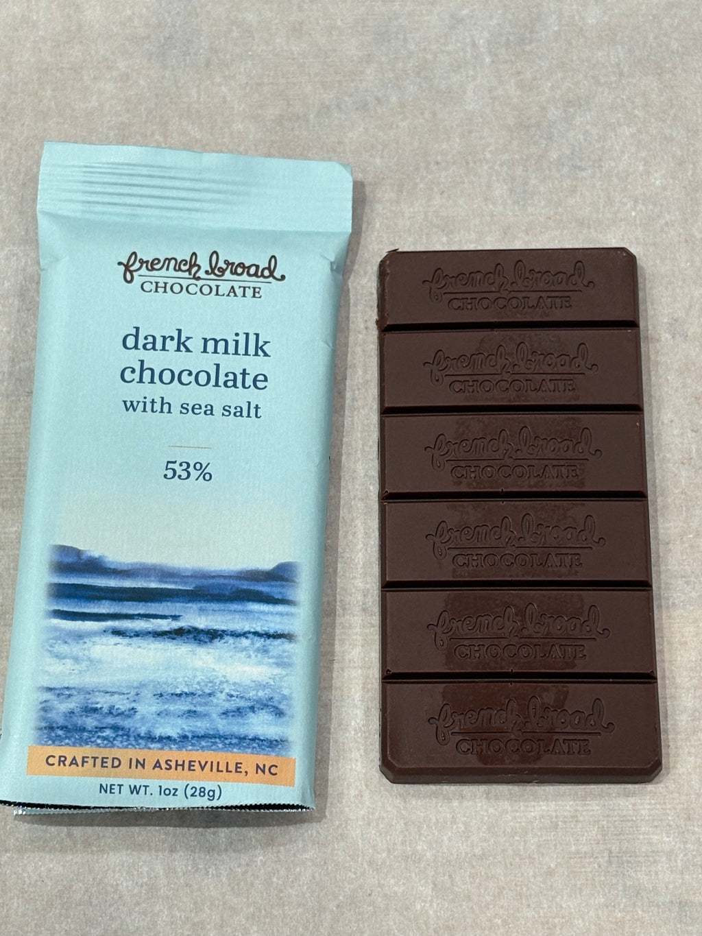 French Broad Chocolate 53% Dark Milk Chocolate Bar with Sea Salt unwrapped showing embossed segments next to light blue packaging