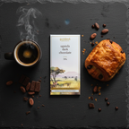 French Broad Chocolate 70% Uganda Dark Chocolate bar in white and gold packaging standing upright on dark marble beside a steaming espresso and flaky croissant