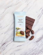 French Broad Chocolate 70% Uganda Dark Chocolate bar and light blue wrapper with cacao beans on white marble surface, crafted in Asheville NC, 1oz