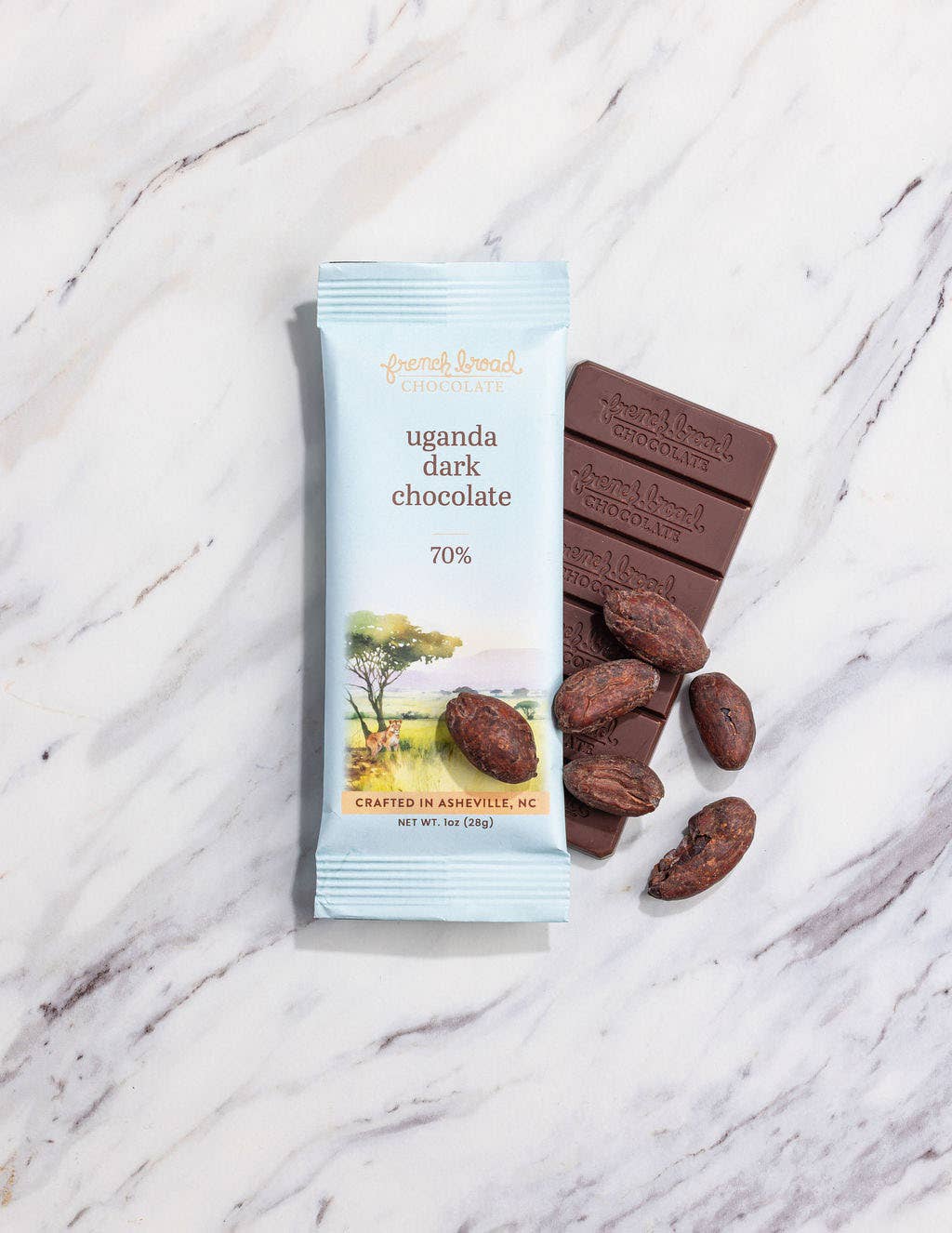 French Broad Chocolate 70% Uganda Dark Chocolate bar and light blue wrapper with cacao beans on white marble surface, crafted in Asheville NC, 1oz