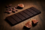 French Broad Chocolate 70% Uganda Dark Chocolate bar on wooden surface with cacao beans and figs around the bar.