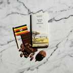 French Broad Chocolate 70% Uganda Dark Chocolate bar in white and gold packaging standing upright on warm wood with a Uganda flag, cacao beans, and dried fig