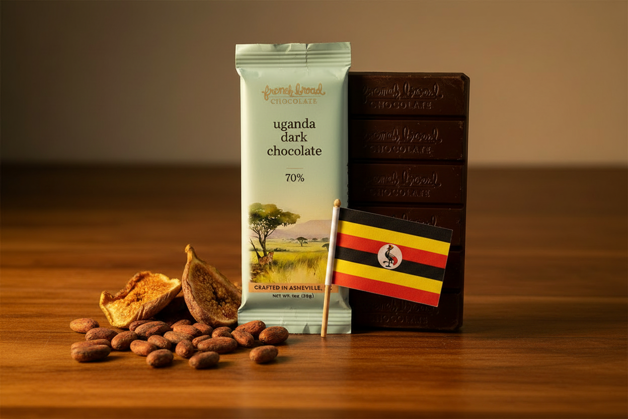 French Broad Chocolate 70% Uganda dark chocolate bar in light blue packaging on dark wooden surface with raw cacao beans and Uganda flag, dramatic side lighting