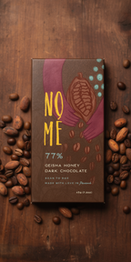 NOME 77% Geisha Honey Dark Chocolate bar surrounded by coffee beans on a wooden surface