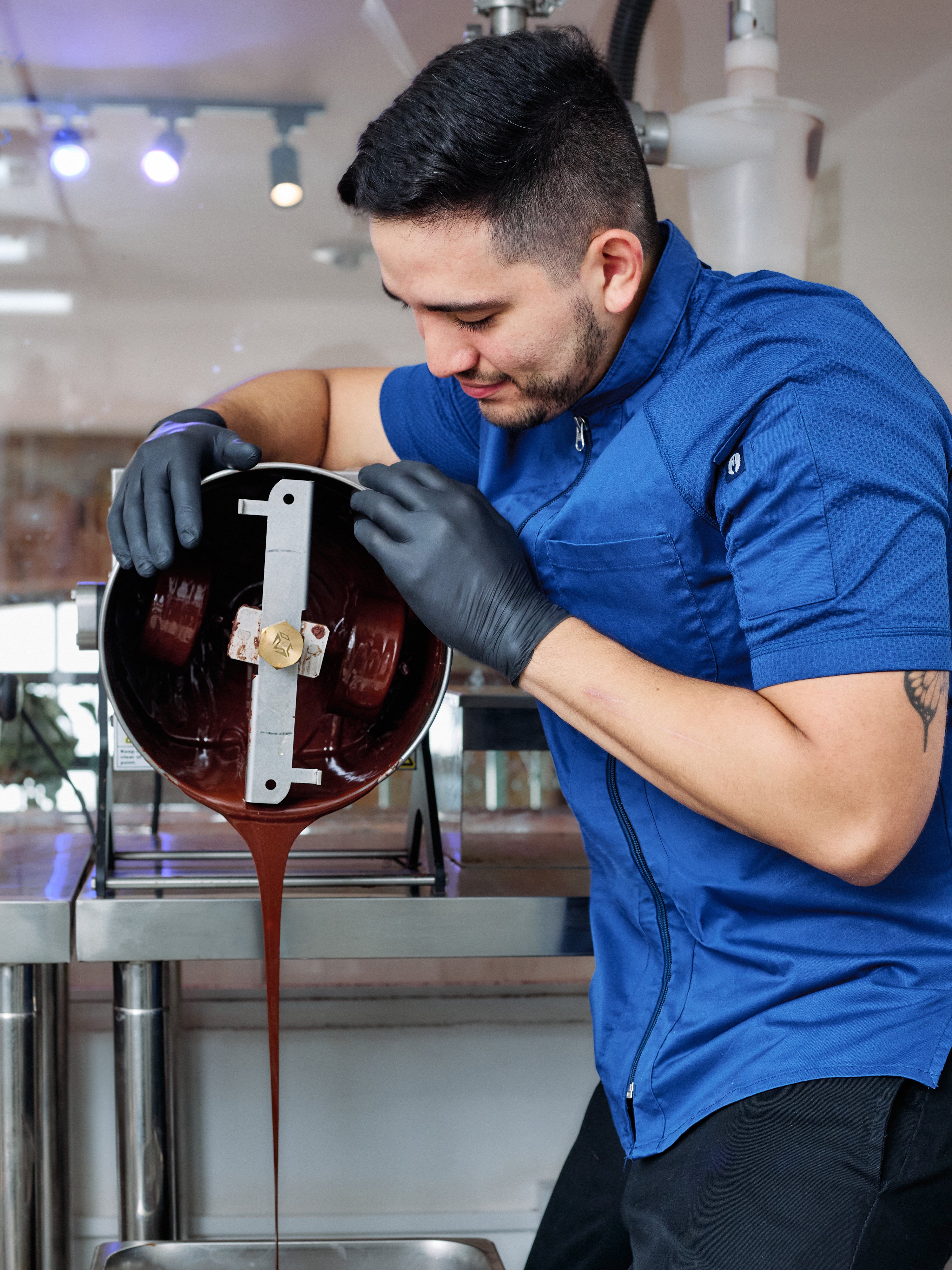 A man in a blue chef's jacket and black gloves pours dark chocolate from a metal bowl into a container in a kitchen setting.