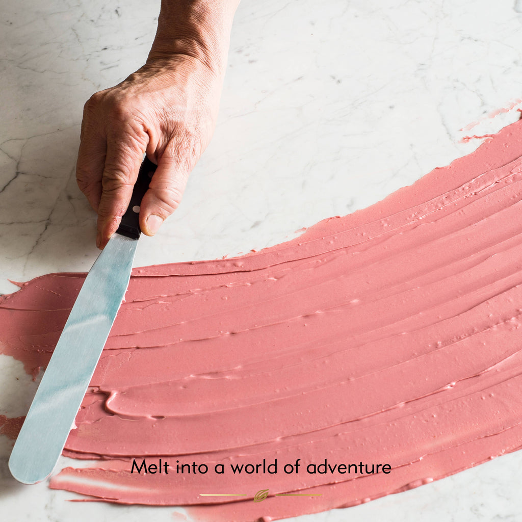 Hand using a spatula to spread pink candy on a white surface with text 'Melt into a world of adventure'.