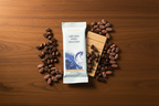 Overhead flat lay of French Broad Chocolate Café Latte White Chocolate bar in light blue packaging on a warm dark wooden table surrounded by roasted coffee beans and raw cacao beans