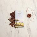 French Broad Chocolate Uganda Dark Chocolate 70% bar on white marble with raw cacao beans, dried fig, and coconut flake representing tasting notes of chocolate cake, fig, and coconut, single-origin Semuliki Forest Uganda