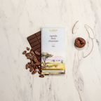 French Broad Chocolate Uganda Dark Chocolate 70% bar on white marble with raw cacao beans, dried fig, and coconut flake representing tasting notes of chocolate cake, fig, and coconut, single-origin Semuliki Forest Uganda