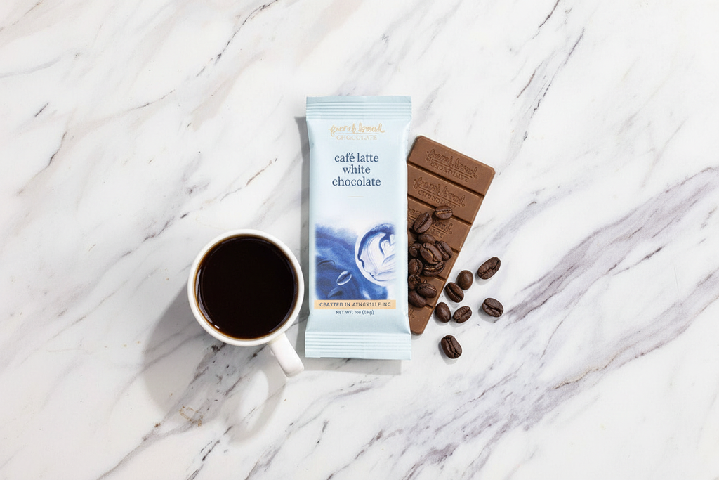 French Broad Chocolate Café Latte White Chocolate bar in light blue packaging styled on a warm marble surface with scattered roasted coffee beans and a small ceramic espresso cup, soft natural light