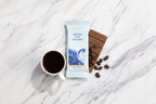 French Broad Chocolate Café Latte White Chocolate bar in light blue packaging styled on a warm marble surface with scattered roasted coffee beans and a small ceramic espresso cup, soft natural light